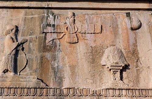 Exemple typique de stèle ornant le haut des tombes des rois achéménides. En haut, à côté du soleil, on peut reconnaître le signe d'<i>Ahura Mazda</i>, Dieu du feu et du soleil qui veille sur le défunt Roi.